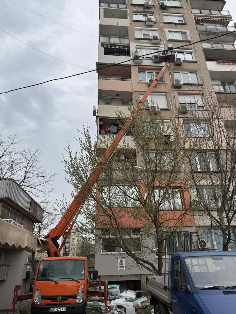 Автовишка със стрела, вдигната между дървета и сграда